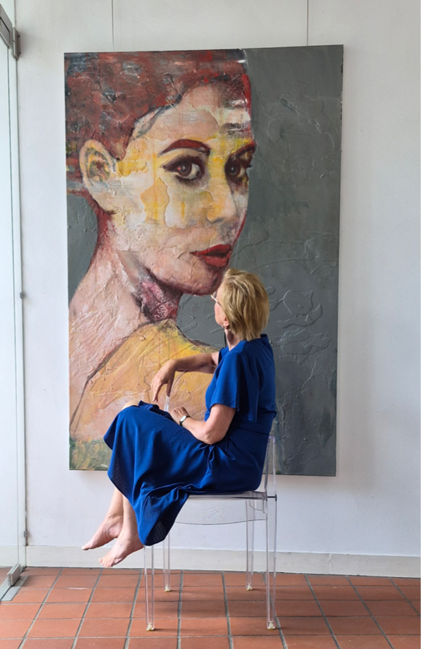 Marian Siereveld in een blauwe jurk, zittend voor haar schilderij in mijn atelier